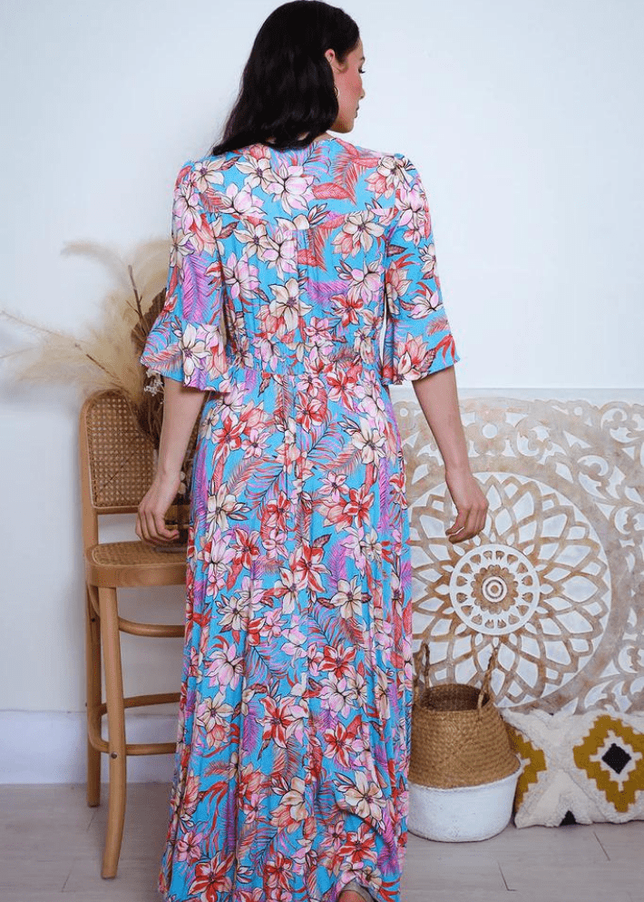 A long, flowing maxi dress adorned with a tropical floral pattern in bright hues of blue, pink, and orange, set against a sky-blue background. The dress features a V-neckline, short flutter sleeves, and a tiered skirt. Brand Avamia