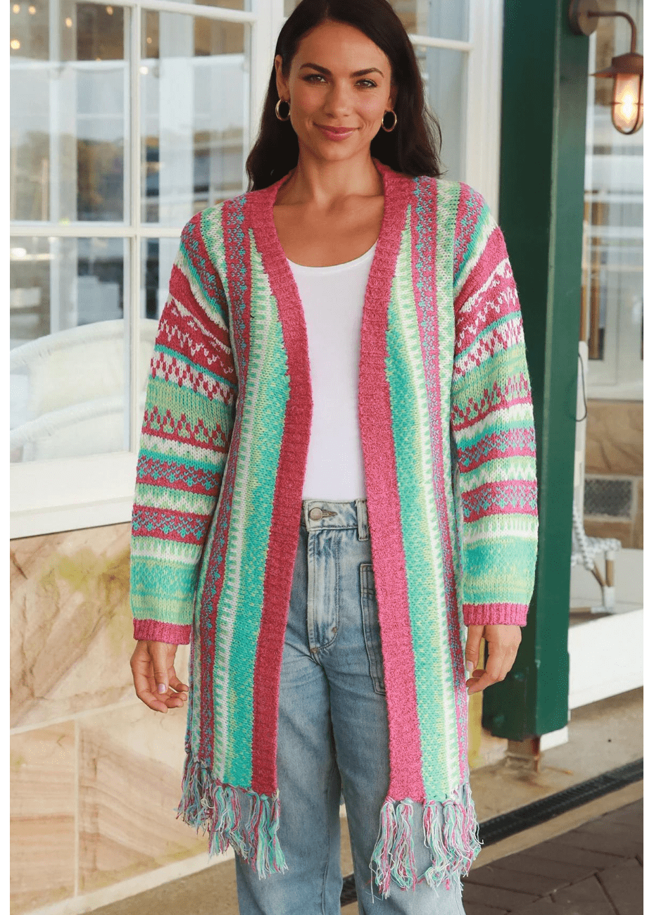 A woman wearing a long, open-front knit cardigan with colourful, geometric patterns in shades of green, pink, white, and blue. The boho cardigan has fringes at the hem and is paired with a white top and high-waisted blue jeans.