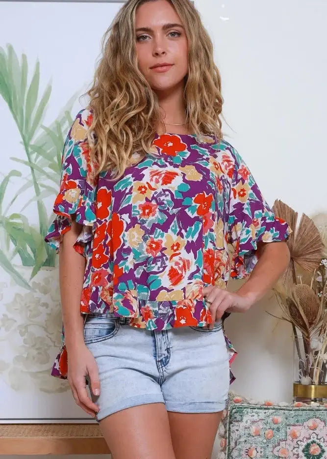 Loose-fitting blouse with a bold floral print in vivid colors of red, orange, purple, and turquoise. The blouse has a round neckline, elbow-length sleeves with a slight ruffle, and a peplum hem. Brand Avamia