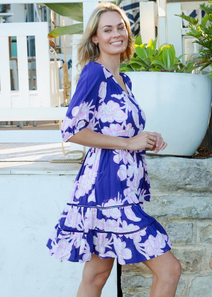 A woman wearing a blue mini dress with oversized pink floral print. A flattering neckline that adds a touch of sophistication. Short sleeves with a puffed design for a stylish look. matching fabric belt to accentuate your waistline. Made from soft, breathable material, perfect for warm summer weather.