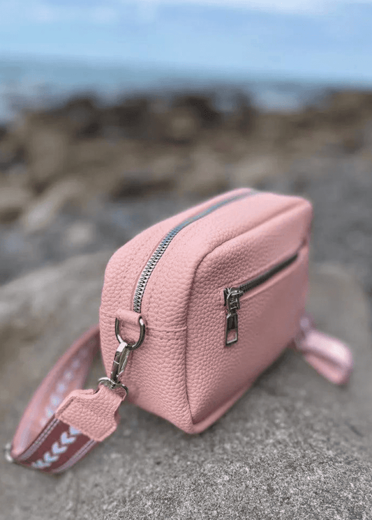 A pink crossbody bag made of vegan leather with a textured surface, featuring silver zippers and hardware. The bag is placed on a rocky surface with a blurred background, showcasing a pink and white, adjustable shoulder strap. 