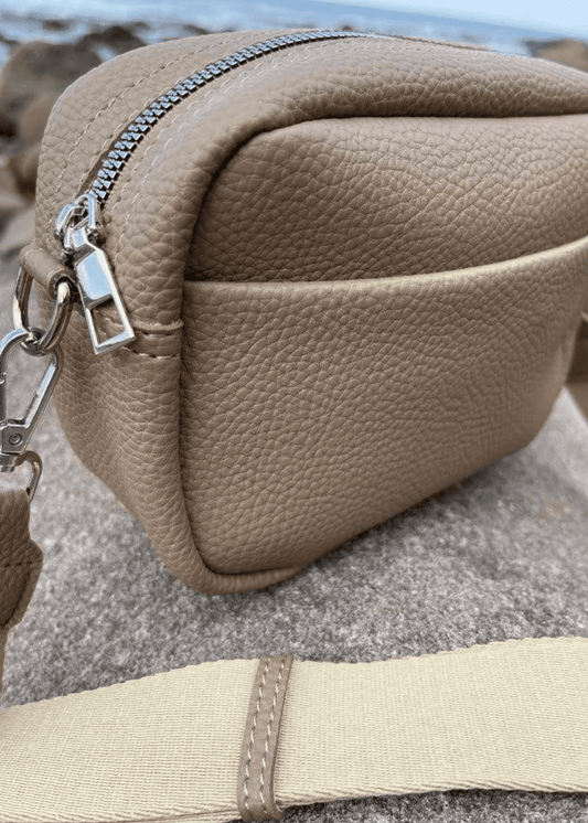 A beige crossbody bag made of vegan leather with a textured surface, featuring silver zippers and hardware. The bag is placed on a rocky surface with a blurred background, showcasing a adjustable shoulder strap. 