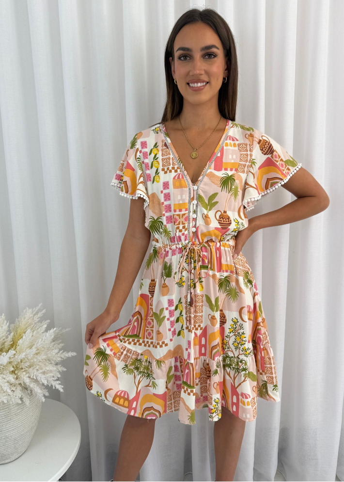 Smiling model wearing an eclectic boho mini dress with architectural motifs, such as domes and arches, alongside natural elements like palm trees and floral accents. The dress showcases a combination of warm tones, including vibrant pink, orange, and green, with a V-neckline, flutter sleeves, an adjustable drawstring waist, and a tiered skirt, perfect for a breezy summer look.