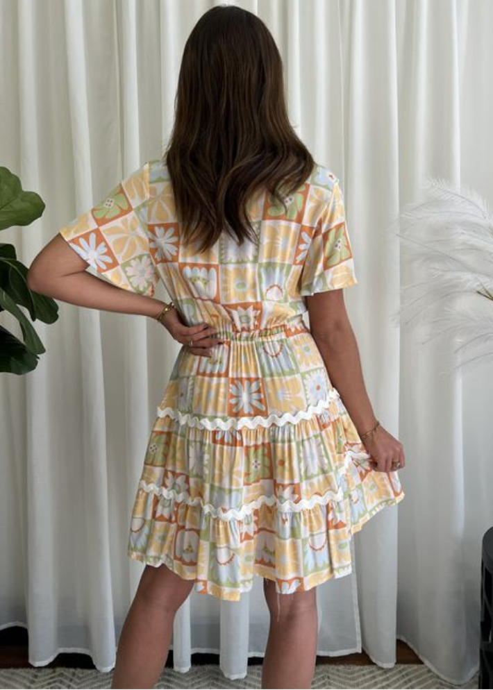 A smiling woman wearing a summer boho-style mini dress with soft yellow, orange, and pastel green floral and geometric patterns. The dress features a V-neckline, fluttery sleeves, an adjustable drawstring waist, and a tiered flared hem with delicate lace detailing, perfect for breezy summer outings.